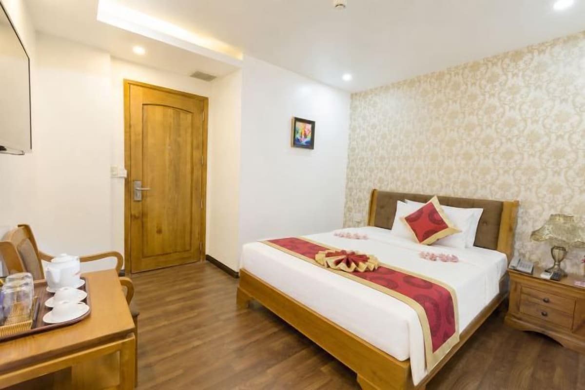 A hotel room with a wooden bed, a bedside table with a lamp, a wooden desk with a chair, and a door.