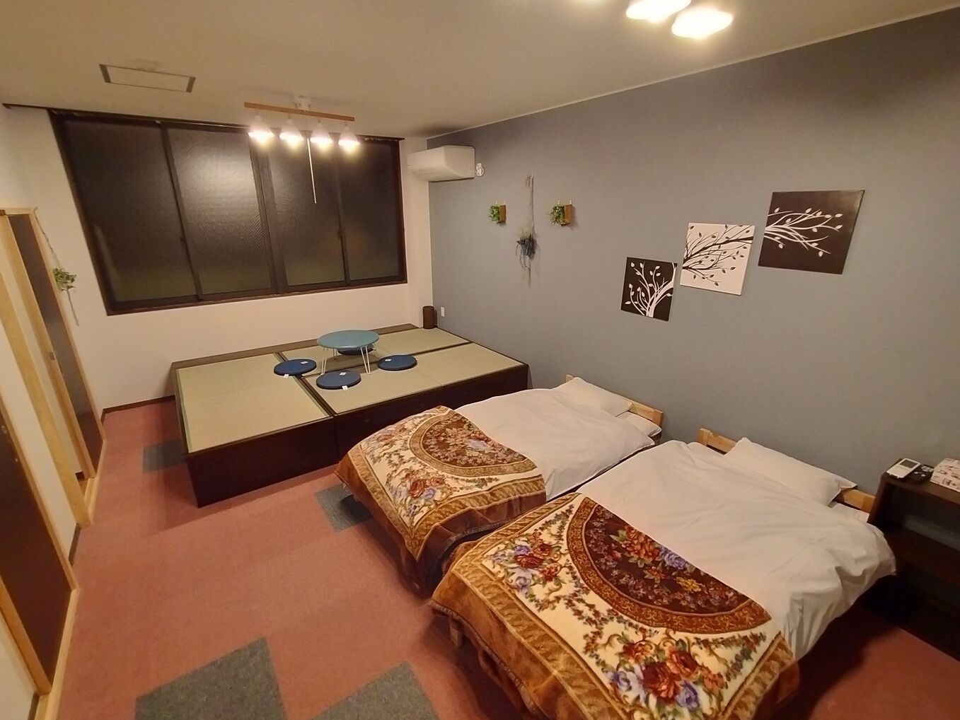 Ein Zimmer mit zwei Betten, einem kleinen Tisch mit Stühlen, einem Fenster mit Jalousien und Wandgestaltungen.
