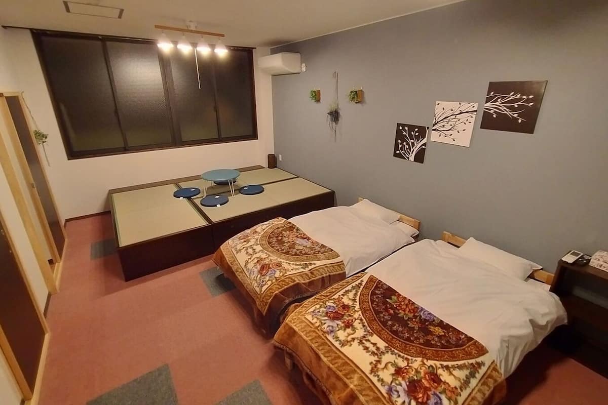 A room with two beds, a small table with chairs, a window with blinds, and wall decorations.
