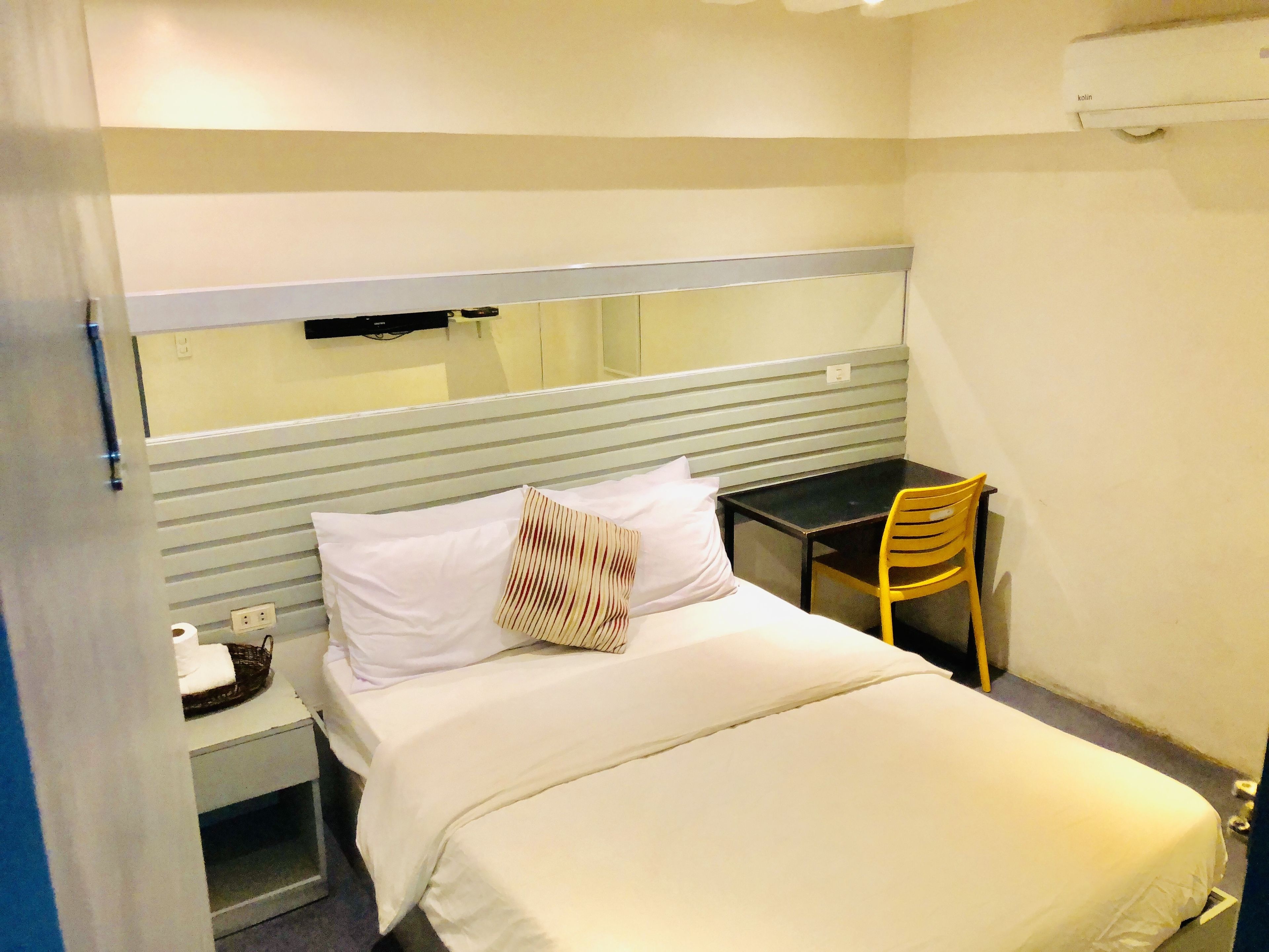 A small, single-bed room with a desk and chair, a mirror above the bed, and a power outlet on the nightstand.