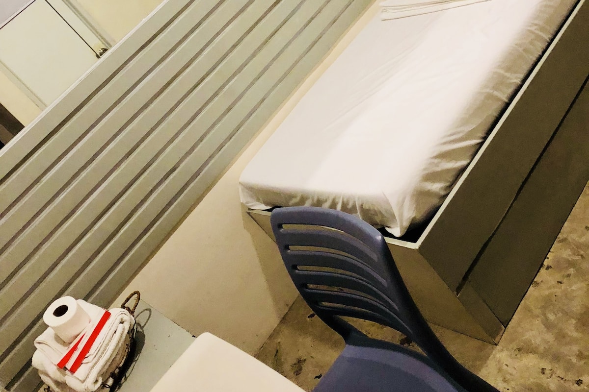 A small, single-bed room with a window, a table with a chair, and a small table with a towel and a roll of toilet paper.