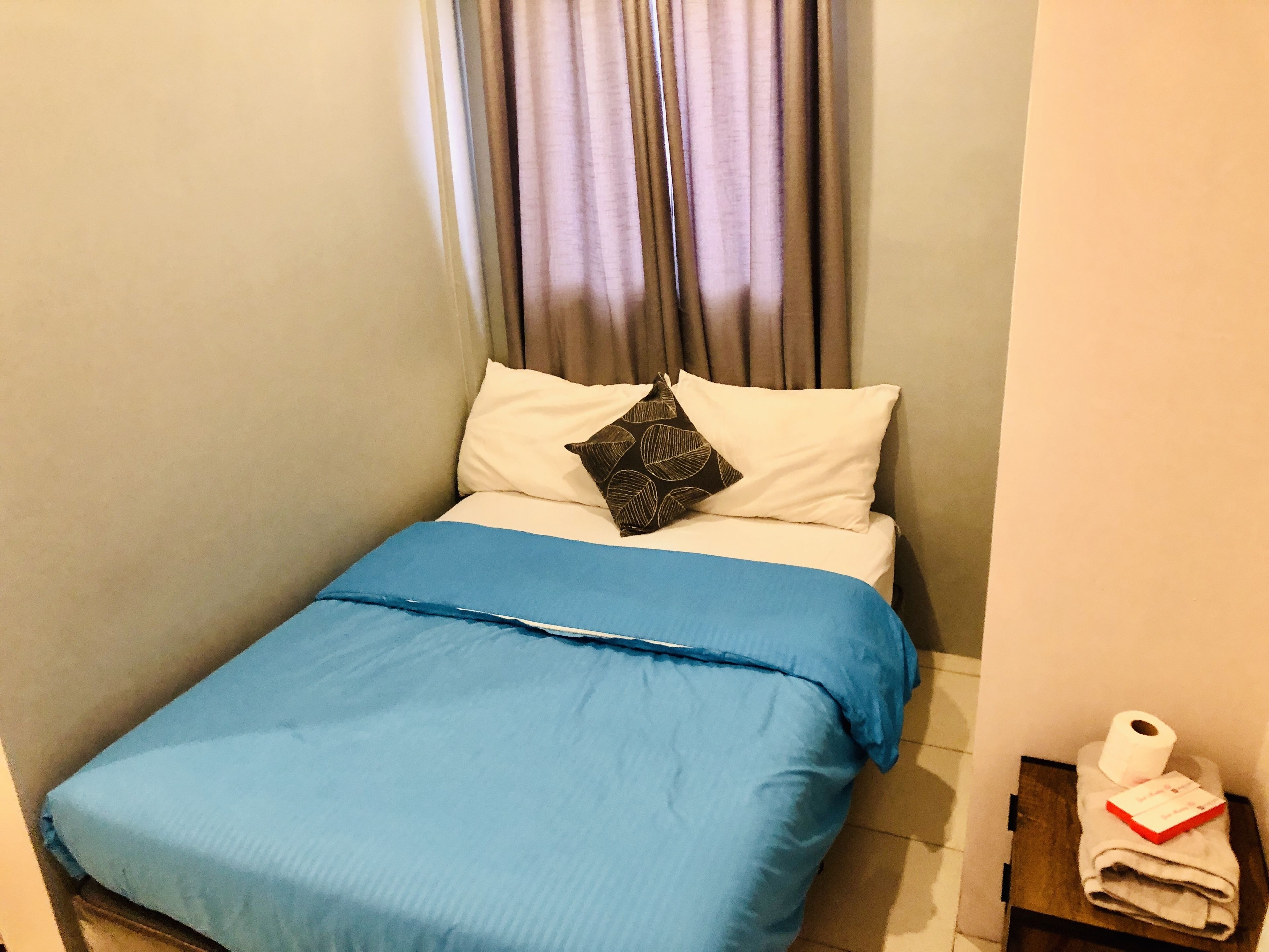 Una cama pequeña y ordenada, con una colcha azul y dos almohadas, una blanca lisa y otra con un estampado de hojas. Se ve una mesita de noche de madera con una toalla y un rollo de papel higiénico a la derecha.