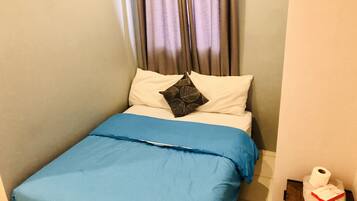 Una cama pequeña y ordenada, con una colcha azul y dos almohadas, una blanca lisa y otra con un estampado de hojas. Se ve una mesita de noche de madera con una toalla y un rollo de papel higiénico a la derecha.