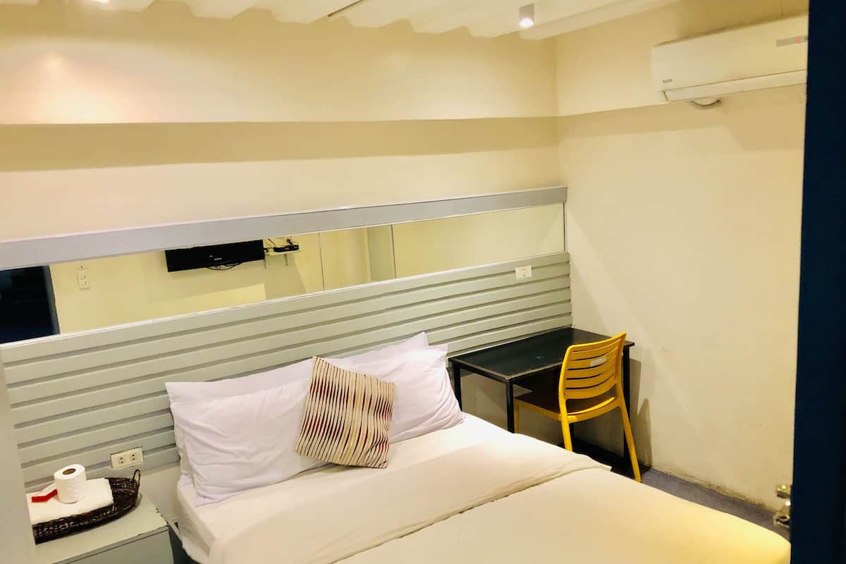 A hotel room with a bed, a desk, a chair, a mirror, and an air conditioning unit.