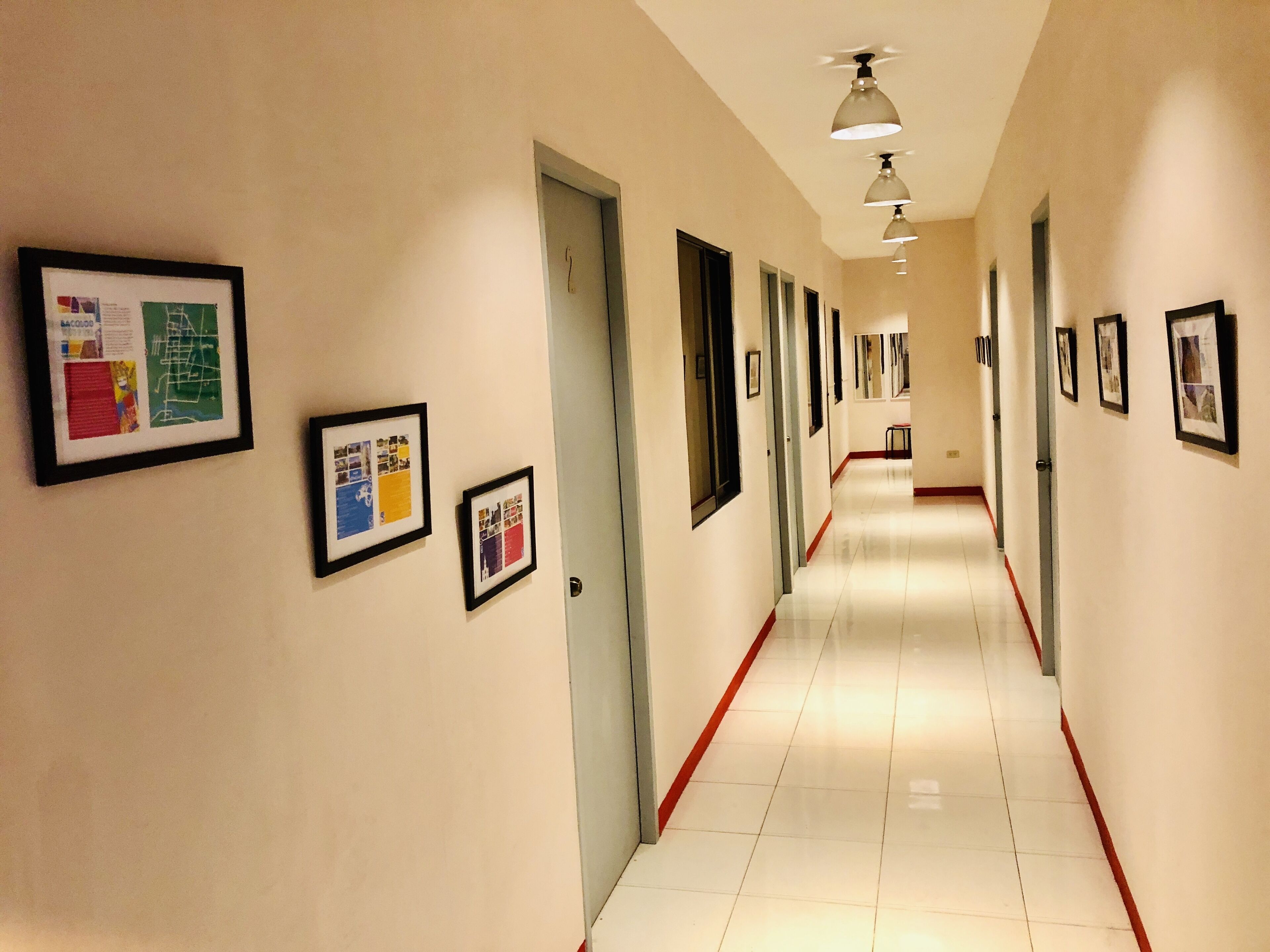 A long, narrow hallway with framed pictures on the walls, a red stripe along the floor, and ceiling lights.