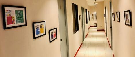 A long, narrow hallway with framed pictures on the walls, a red stripe along the floor, and ceiling lights.