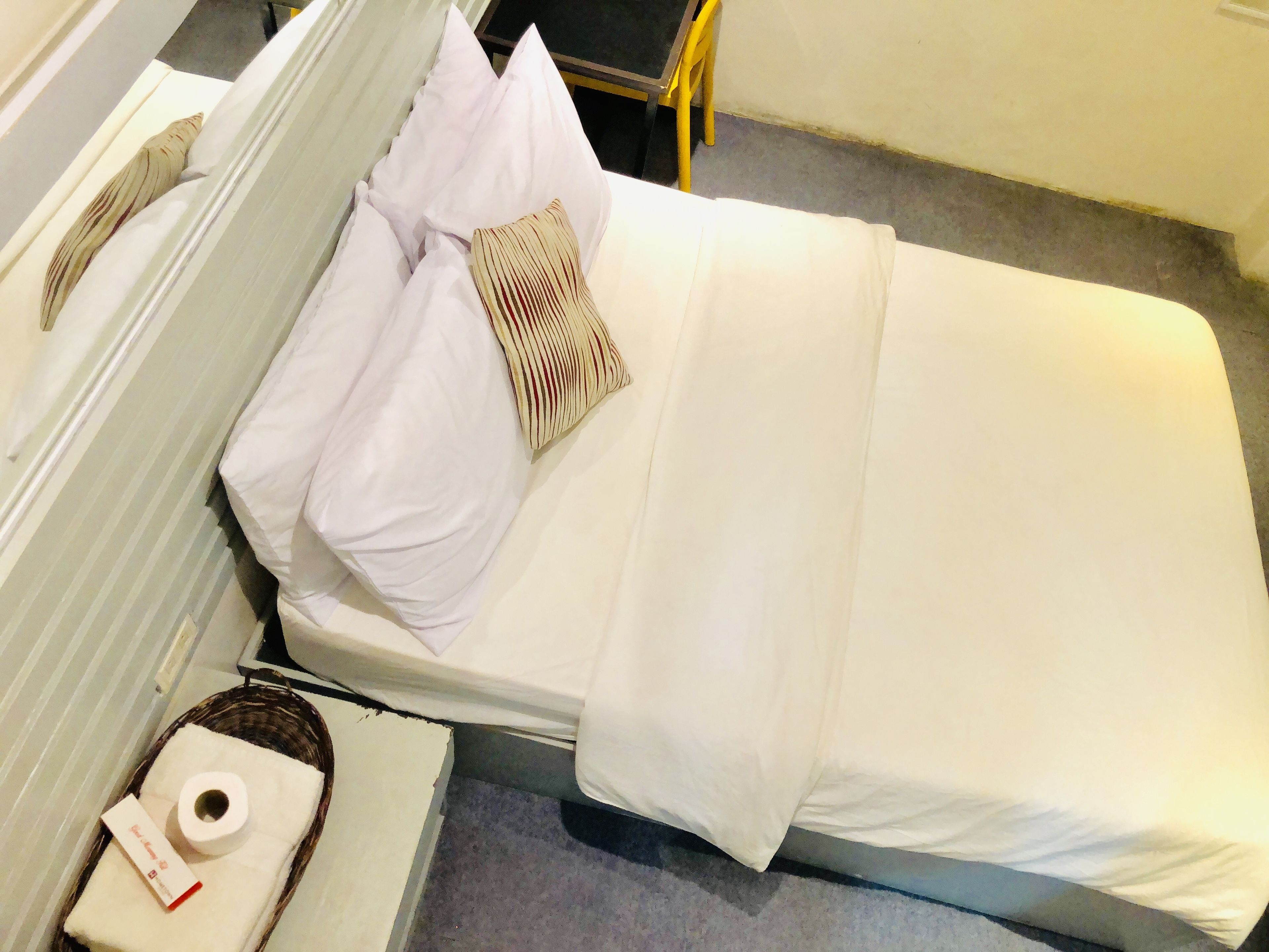 A neatly made bed with white bedding and a striped pillow, a bedside table with a tissue box and a small item, and a chair in the background.
