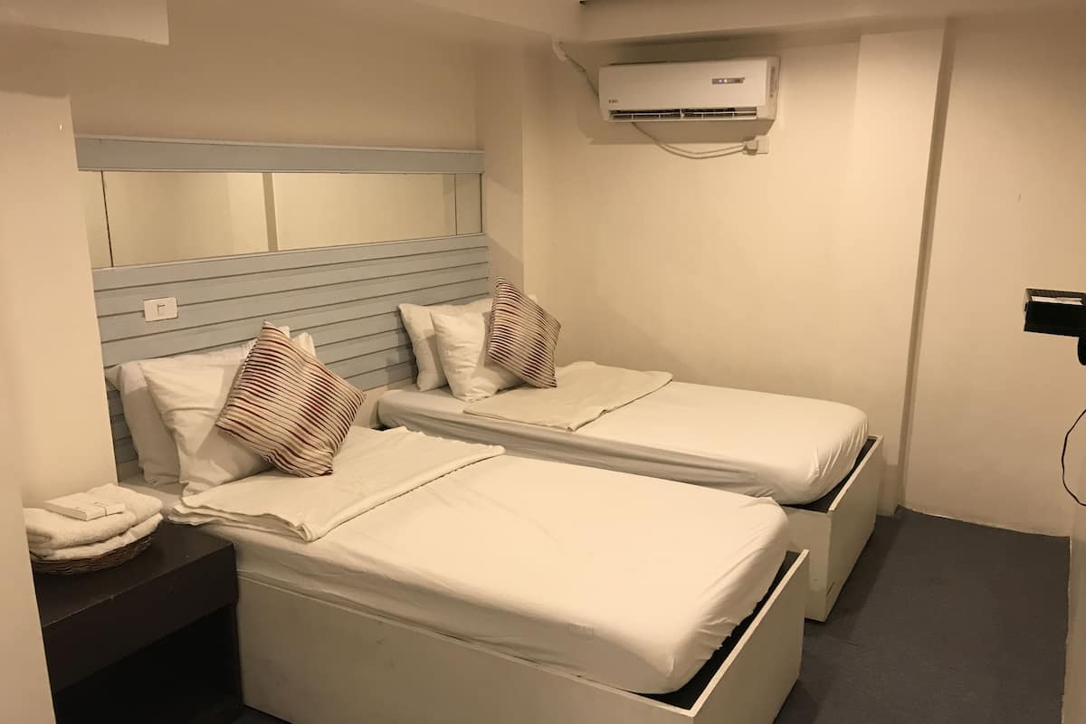 A small, clean room with two single beds, a bedside table, a wall-mounted air conditioner, and a small shelf.