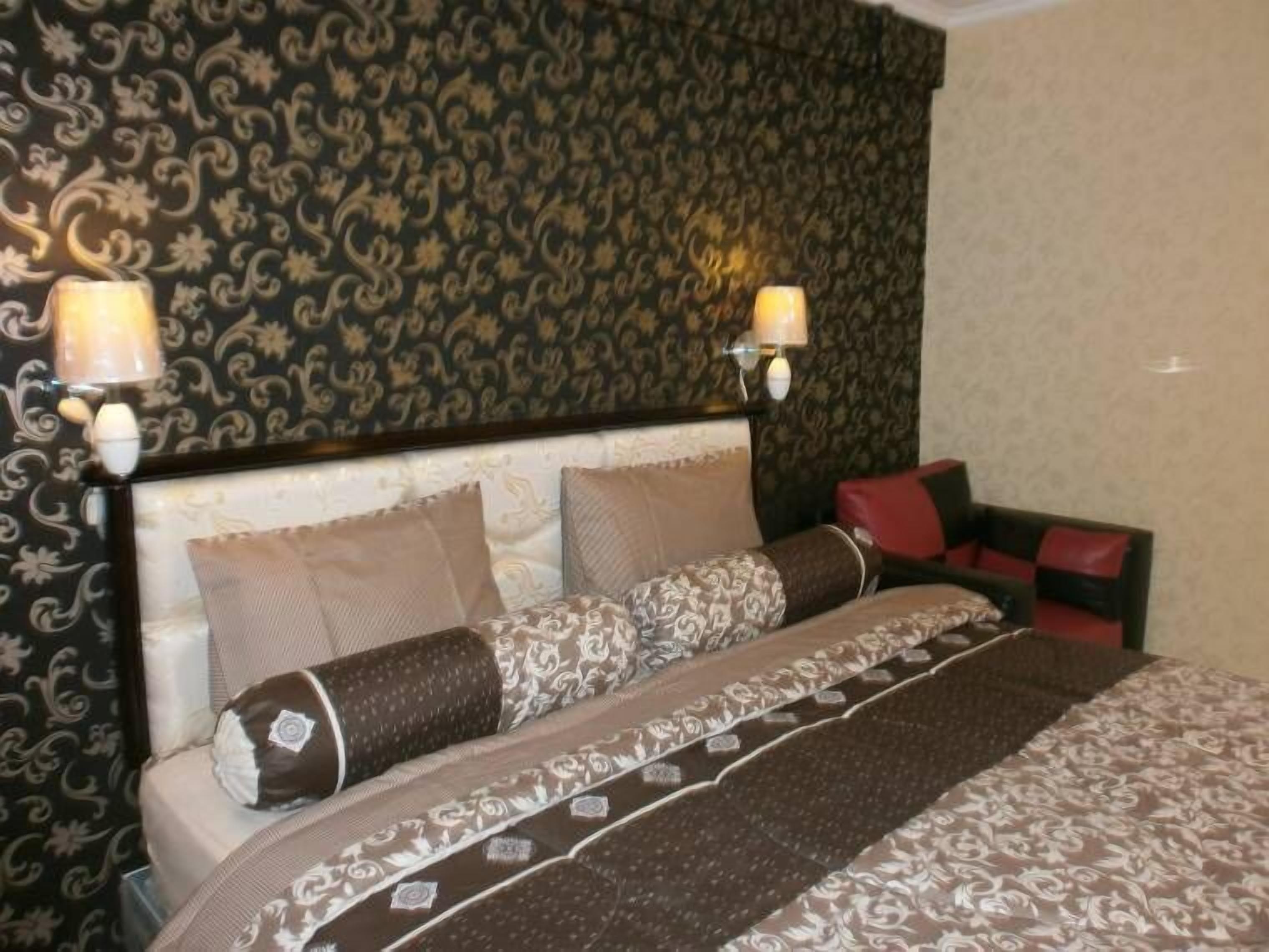 A hotel room with a patterned wallpaper, a bed with multiple pillows, and two wall-mounted lamps.