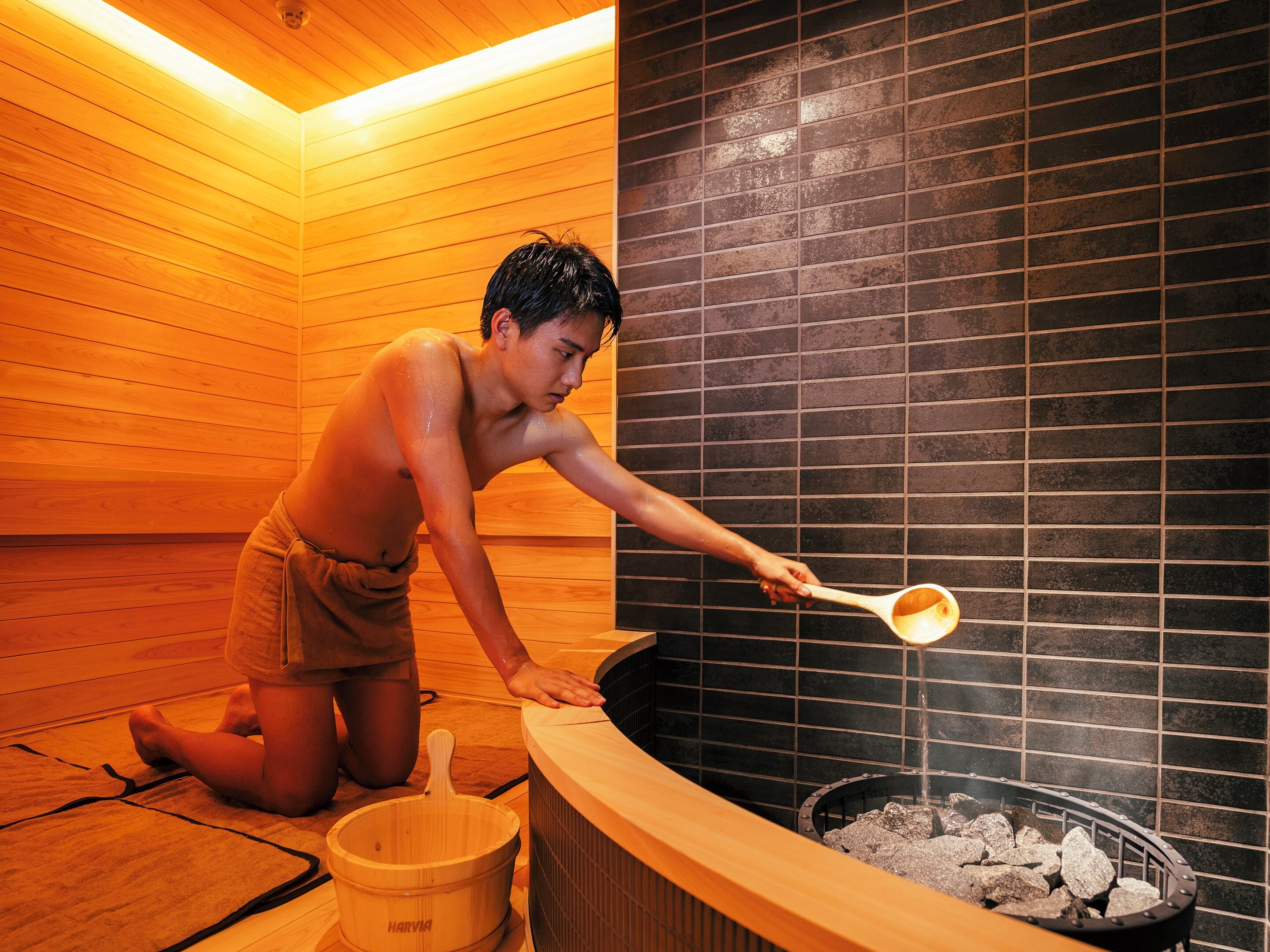 A sauna room with a wooden floor, a black tiled wall, and a wooden tub filled with rocks.
