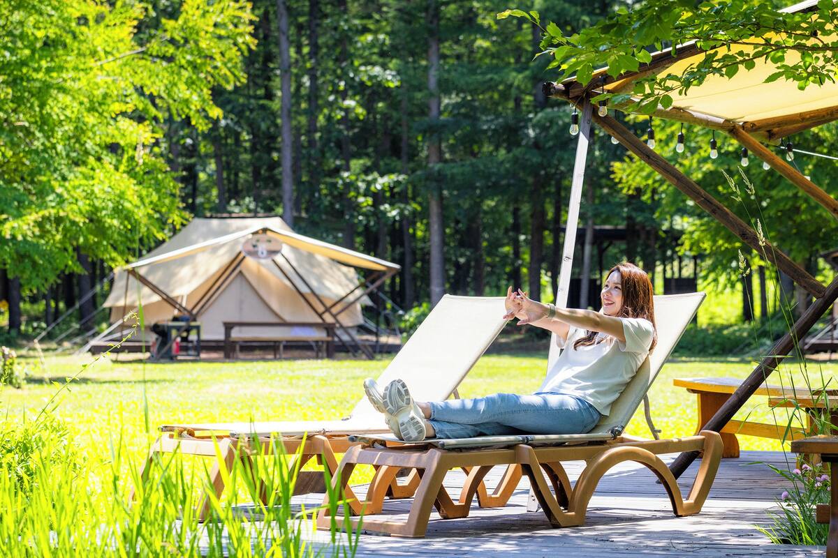 A person relaxing on a wooden lounge chair with a view of a tented accommodation and a wooden deck.