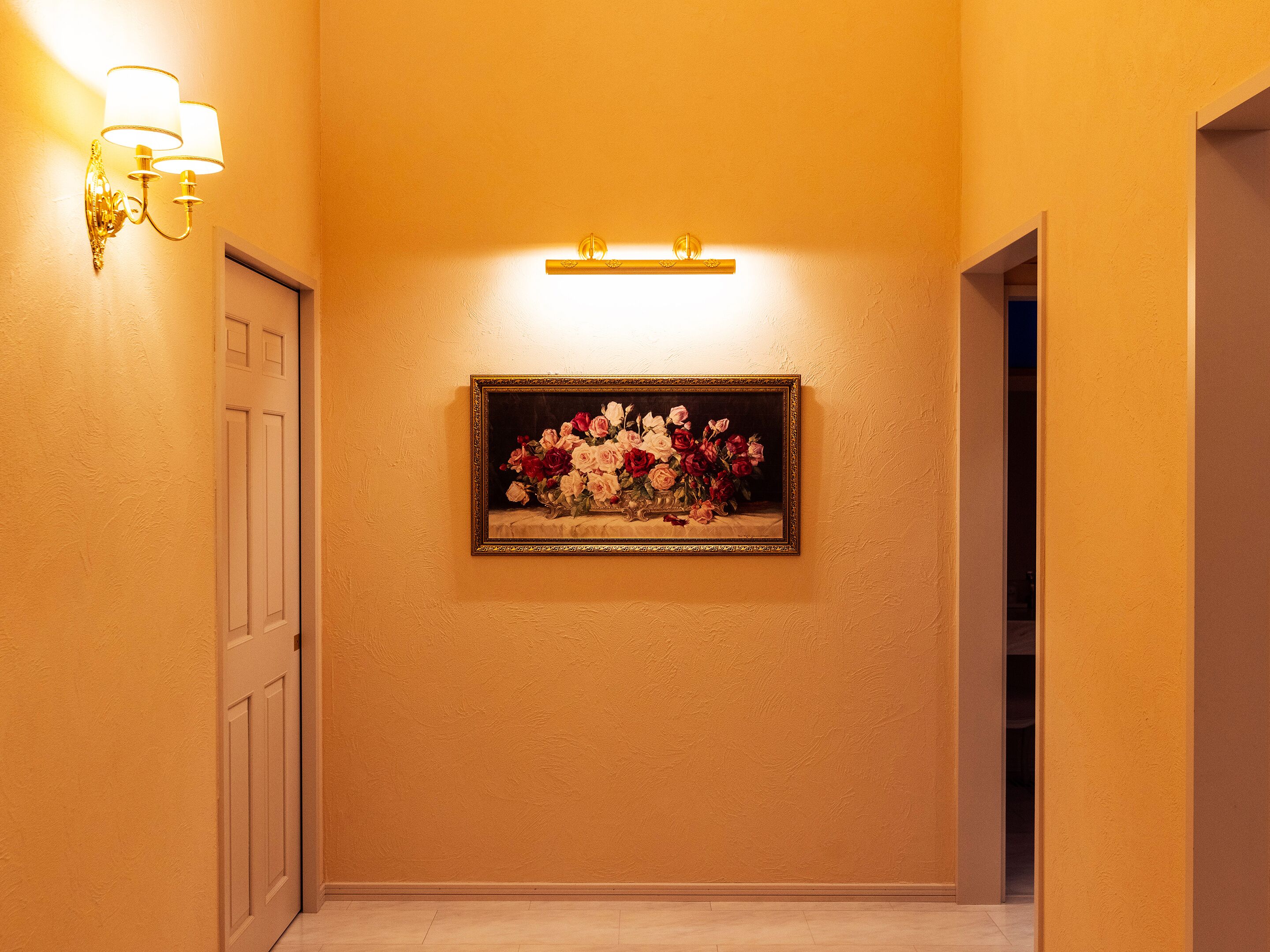 A hallway with a framed floral painting on the wall, a door on the left, and a light fixture above.
