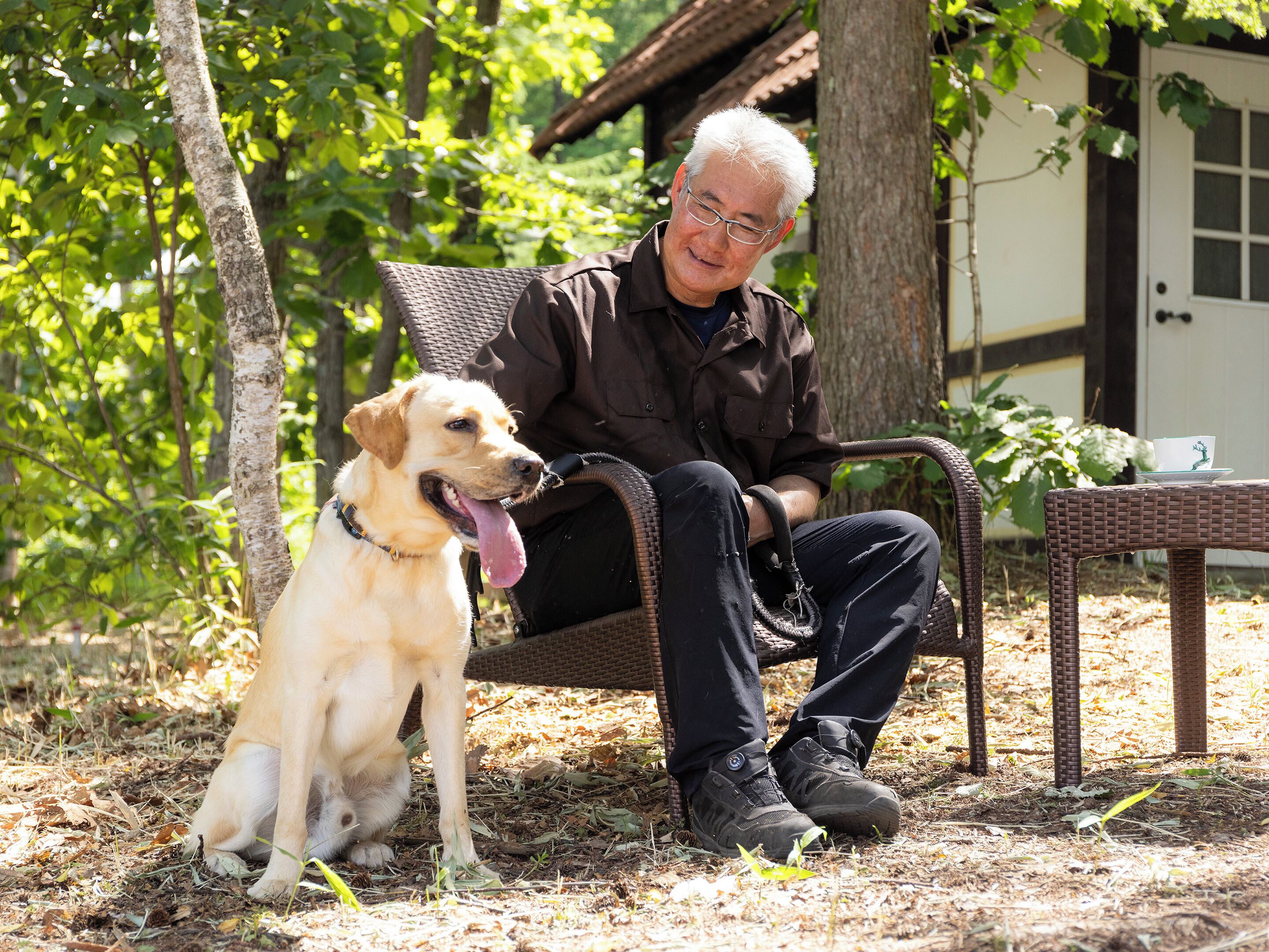An elderly man sitting on a wicker chair outdoors, with a dog beside him.