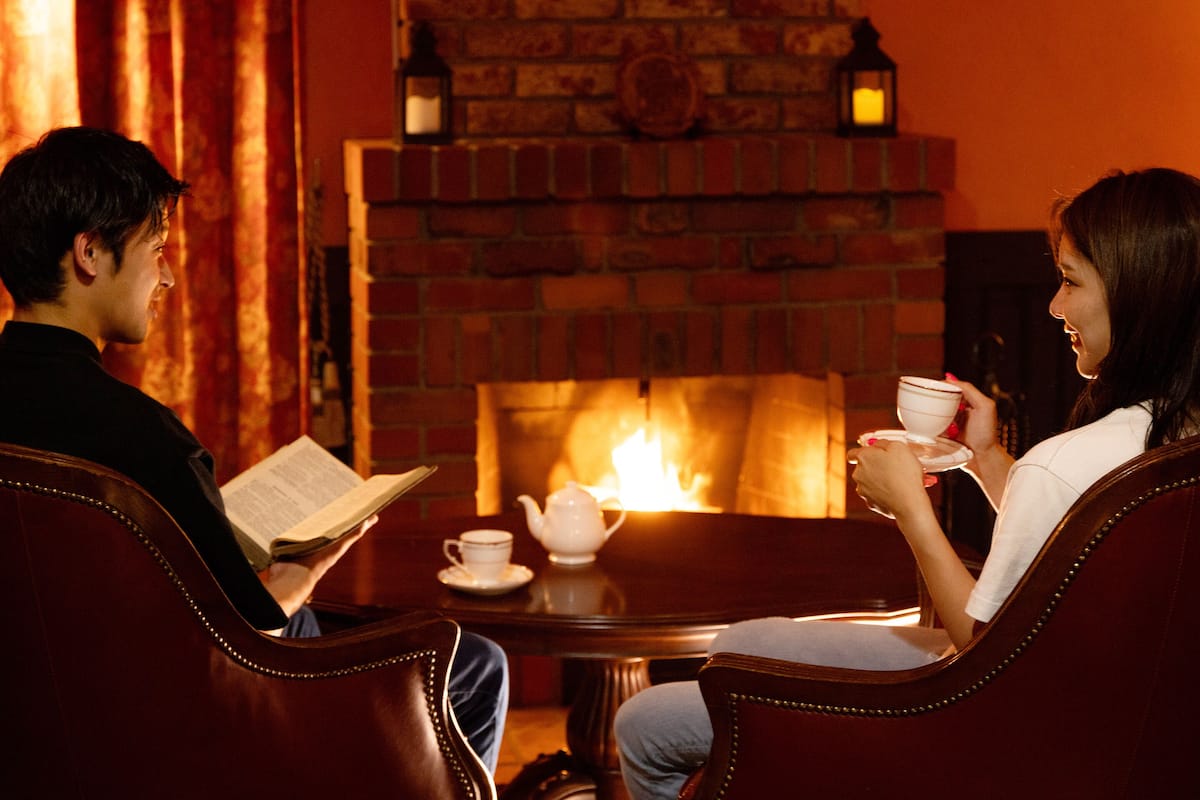 A cozy room with a brick fireplace, leather chairs, a table with a teapot and cups, and a person reading a book.