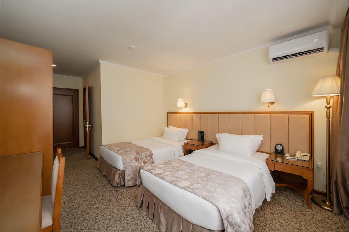 A hotel room with two beds, a desk, a chair, a lamp, and an air conditioning unit.