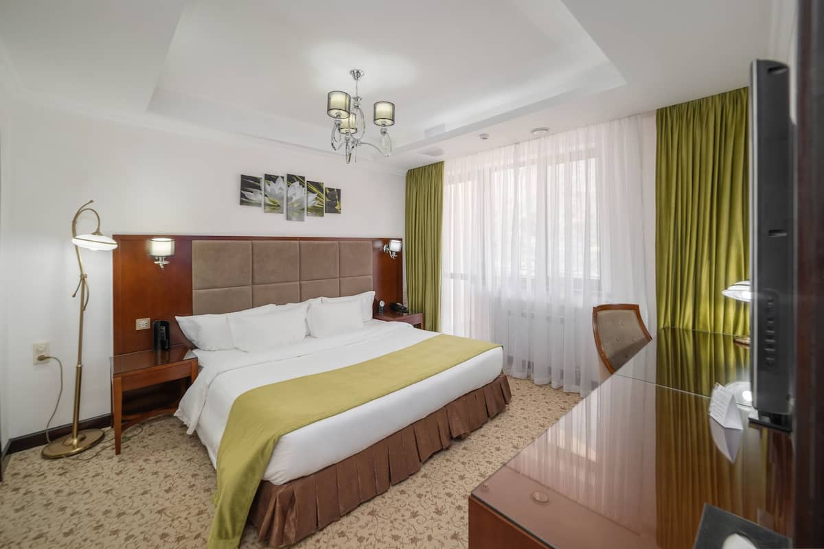 A hotel room with a large bed, a wooden headboard, a desk with a mirror, a floor lamp, and a window with sheer curtains.