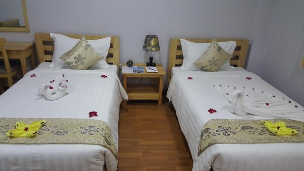 A hotel room with two single beds, each with a wooden headboard, white bedding, and decorative pillows. There is a small wooden nightstand between the beds with a lamp and a book. - Asian Smile Hotel (Yangon)