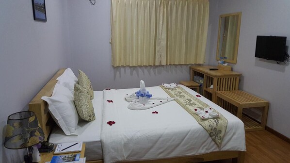 Habitación de hotel con una cama bien hecha, una mesita de noche con lámpara, una mesita auxiliar de madera, un televisor y una ventana con cortinas.