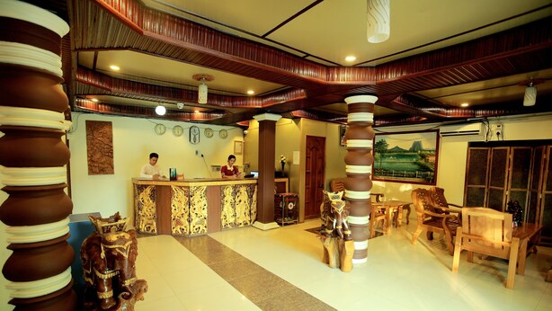 A reception area with a wooden reception desk, a receptionist, and a receptionist's computer. There are wooden chairs and a large wooden sculpture in the background. - Hotel Angel Land (Hpa-An)