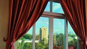 A window with red curtains, a view of a building, and greenery outside. - Hotel Angel Land (Hpa-An)