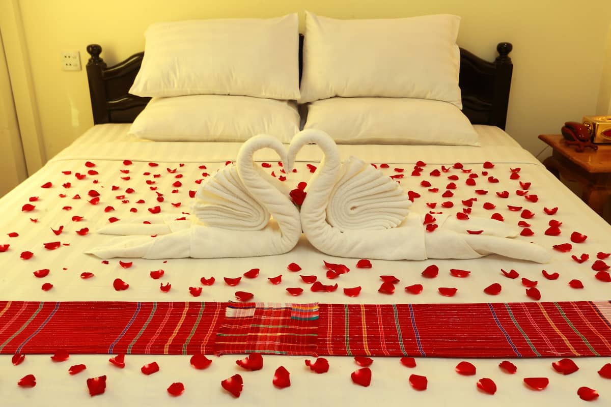 A bed with white linens, a heart-shaped towel arrangement, and red rose petals scattered across the bed.