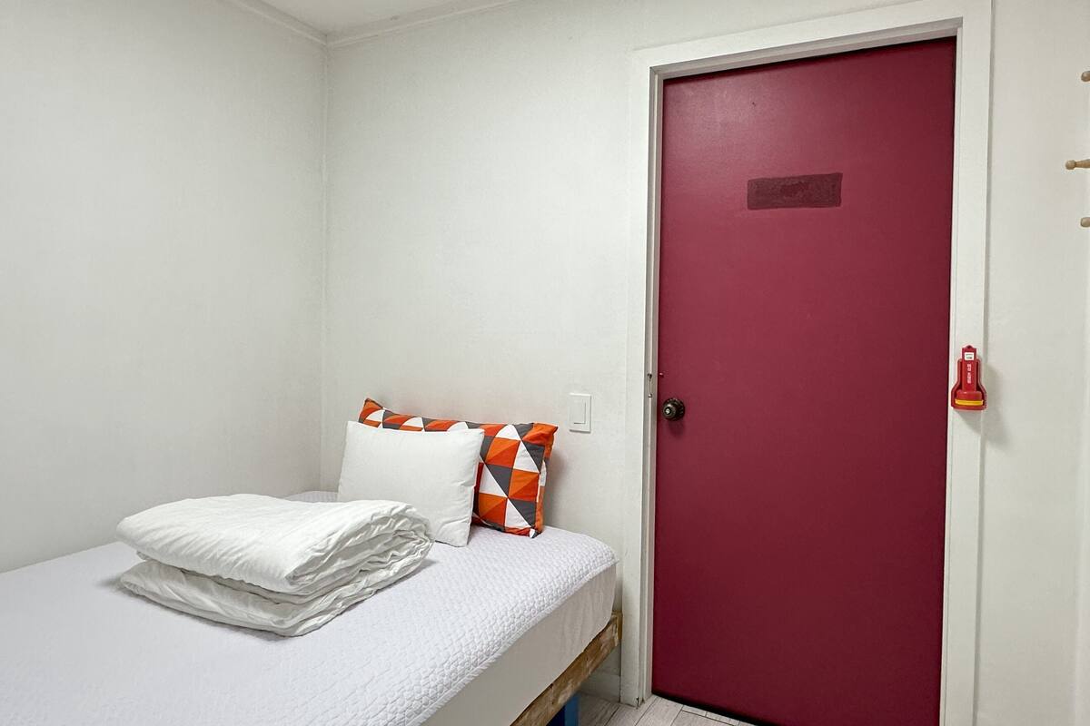 Une petite chambre avec un lit simple, du linge de lit blanc et une porte rouge.