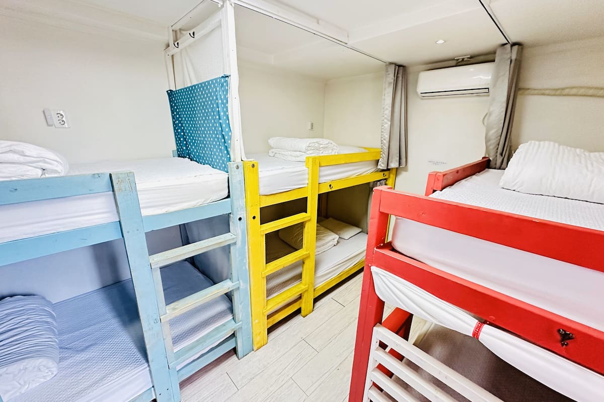 Une chambre de dortoir avec des lits superposés bleus, jaunes et rouges, du linge de lit blanc, un rideau et un appareil de climatisation.