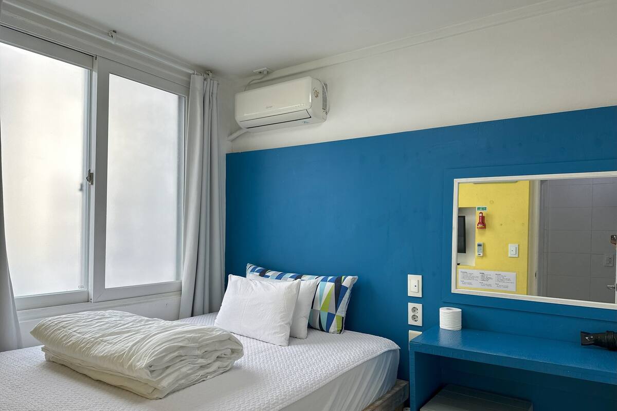 Une chambre d’hôtel avec un lit, une fenêtre avec des rideaux, un appareil de climatisation, un miroir et un mur bleu.