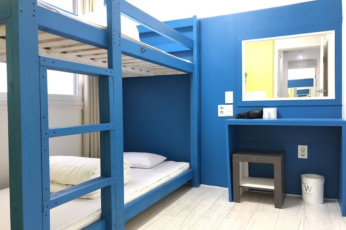 Une chambre avec un lit superposé, un mur bleu, une petite table, une poubelle et un miroir.