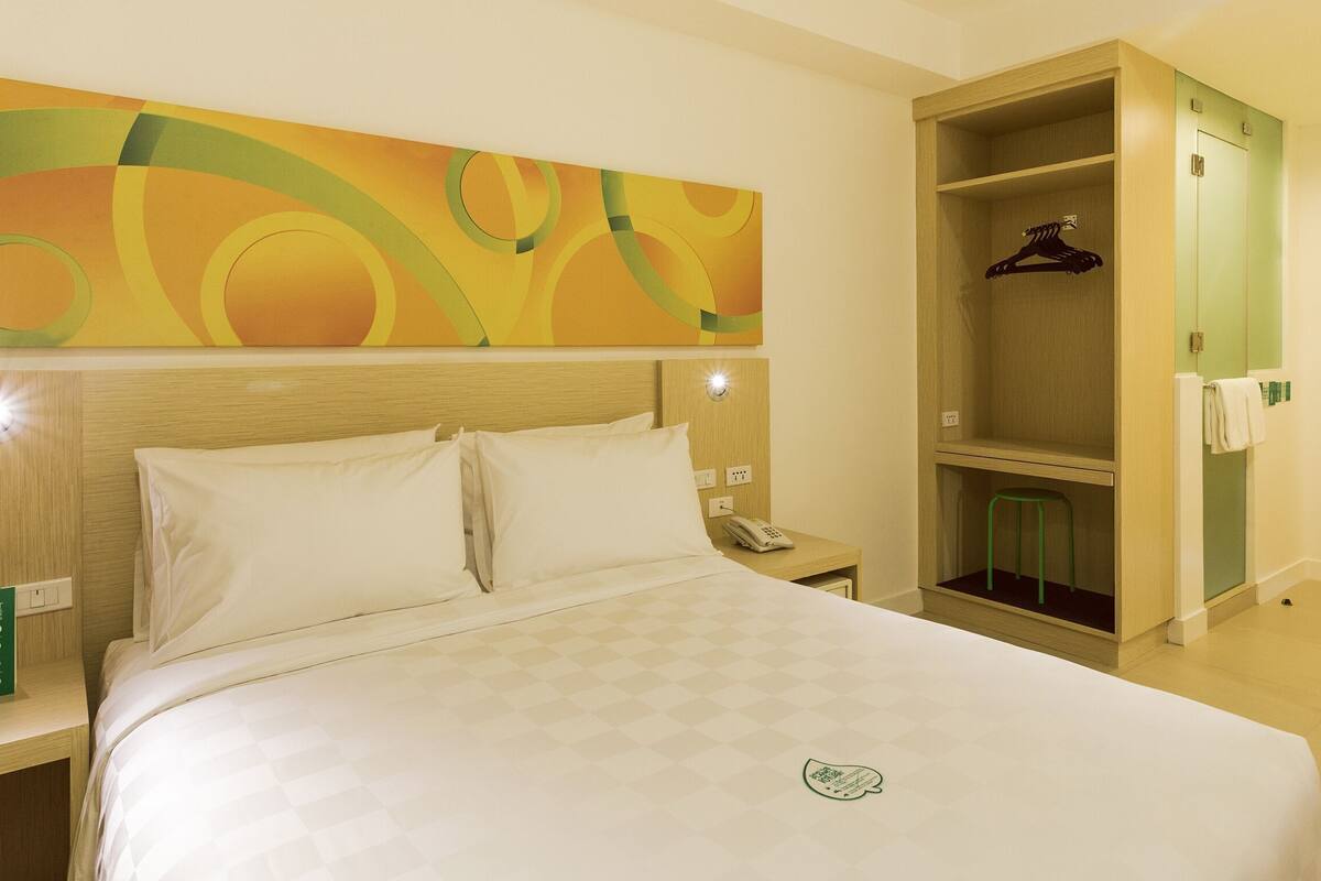 Une chambre d’hôtel comprenant un lit, une table de chevet, un téléphone, un tabouret vert, un porte-manteau et une armoire en bois.