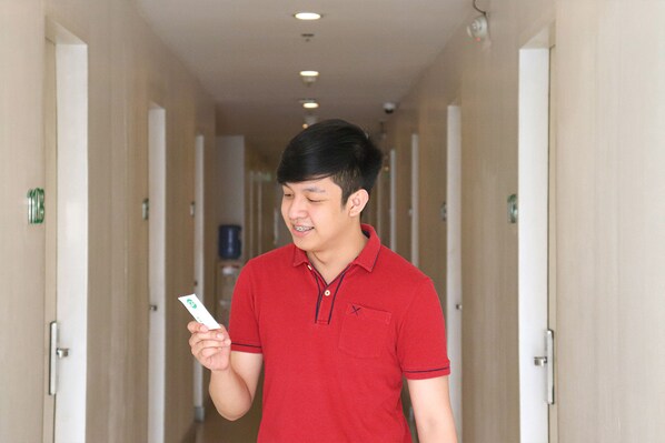 Un hombre con una camisa polo roja sostiene una tarjeta en un pasillo de hotel con paredes y puertas color beige.