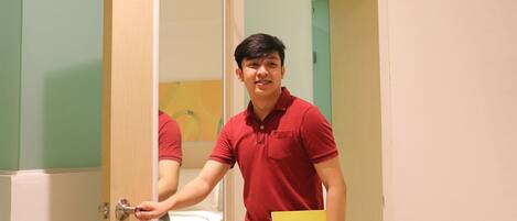 A man in a red polo shirt holding a yellow folder, standing in a doorway with a bed visible in the background.
