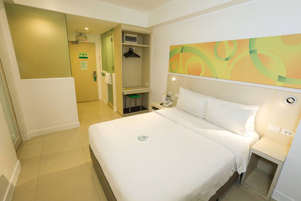 Une chambre d’hôtel avec un grand lit, une table de chevet avec un téléphone, un petit bureau avec une chaise, une armoire et une salle de bain visible à travers une porte ouverte.