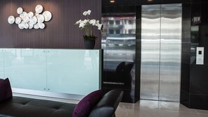 A modern reception area with a glass partition, a sofa with purple cushions, a potted orchid, and a reflection of a building in the glass. - Hotel Mezzo (Kuala Lumpur)