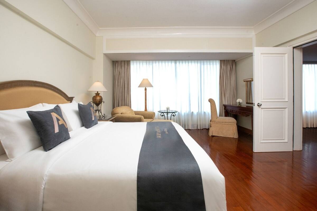 A hotel room with a large bed, a chair, a small table, and a door leading to another room.