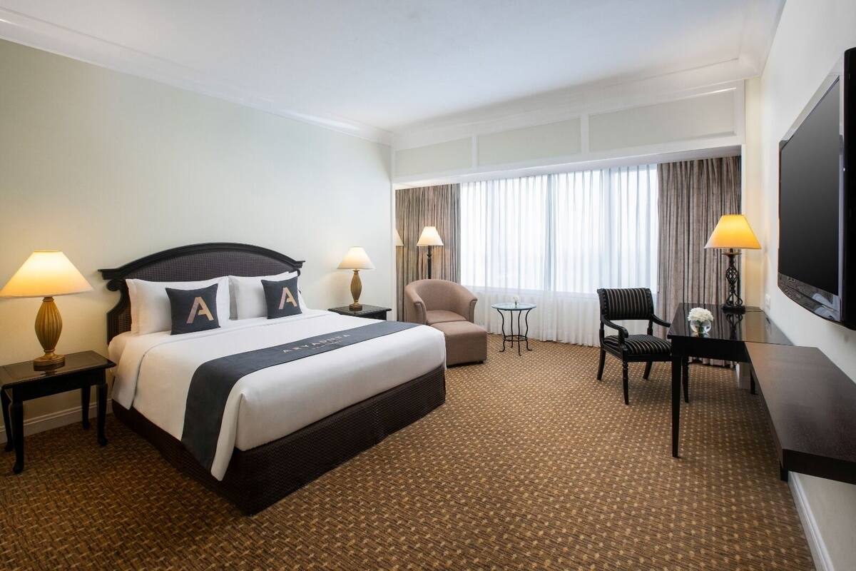 A hotel room with a large bed, two bedside tables with lamps, a sofa, a small table with a flower, and a TV on the wall.
