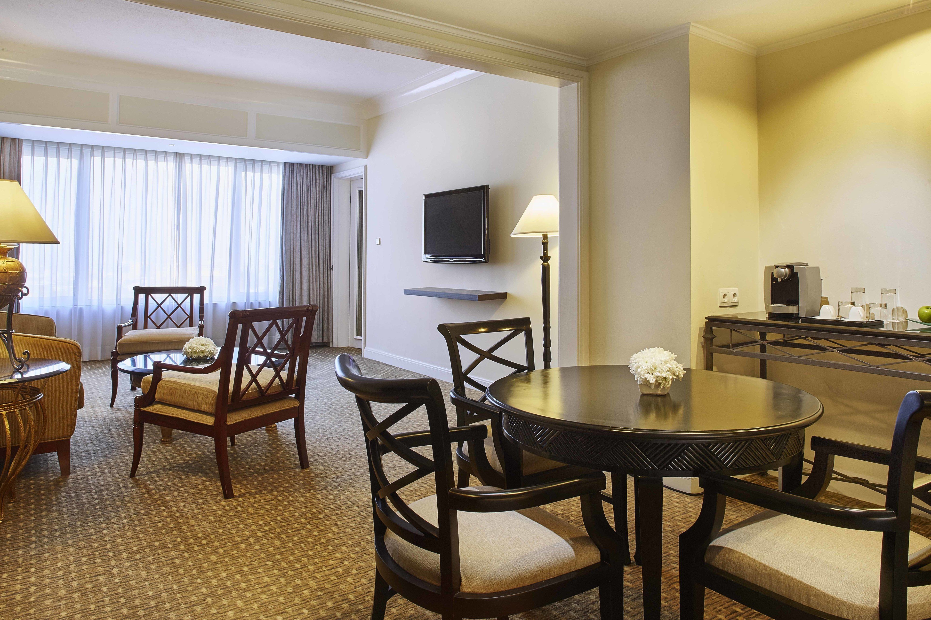 Habitación de hotel con comedor que incluye una mesa redonda, cuatro sillas, un televisor montado en la pared y una cafetera sobre una mesita.