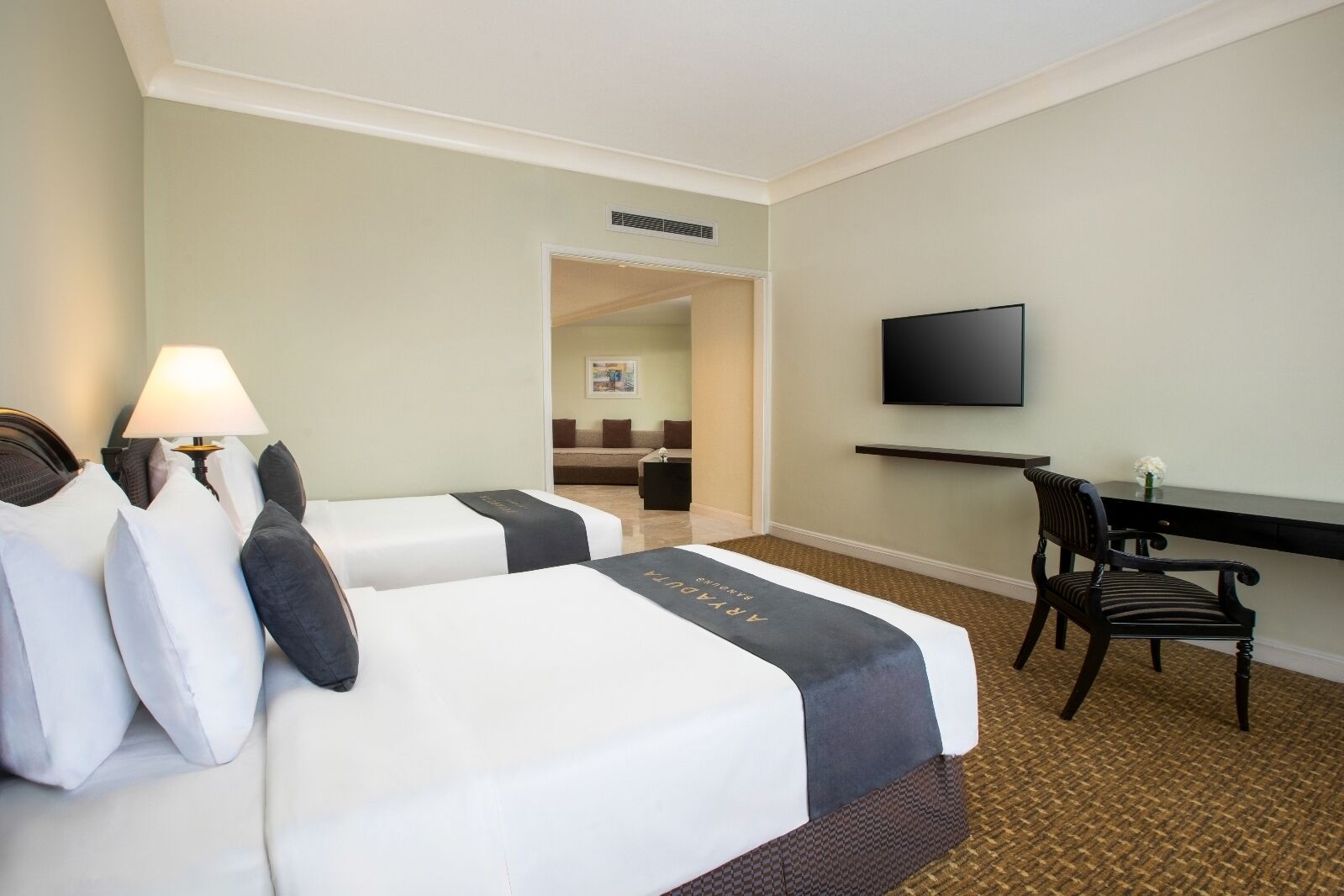 A hotel room with two beds, a desk, a chair, a TV, and a lamp.