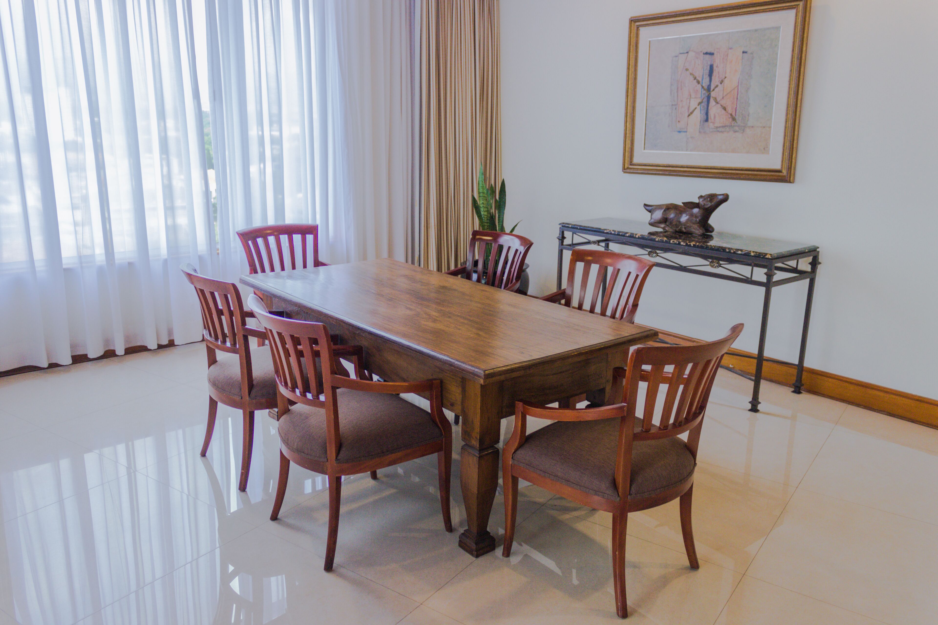 A dining area with a wooden table and six chairs, a potted plant, a framed picture, and a small table with a sculpture.