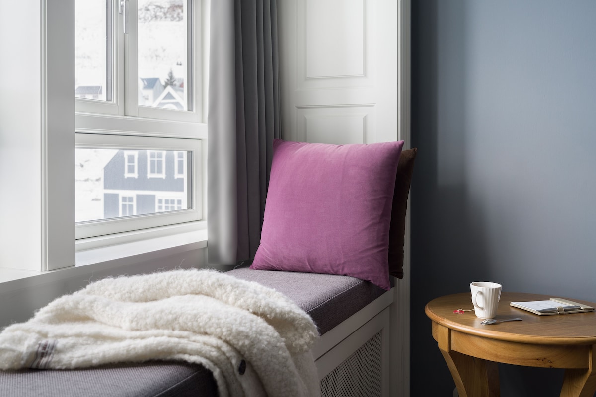A cozy room with a window, a bed with a purple pillow, a white blanket, and a small wooden table with a cup and a spoon.