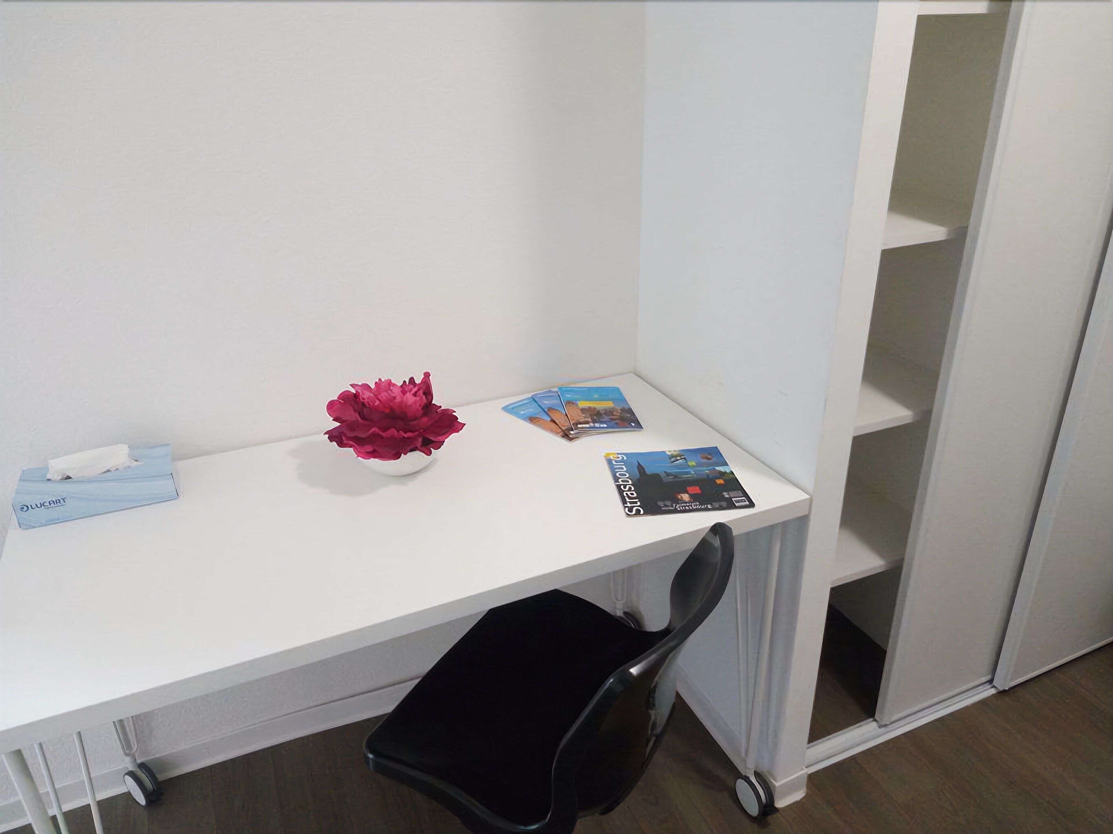 Een wit bureau met een zwarte stoel, een zakdoos, een decoratieve bloem en een paar tijdschriften.
