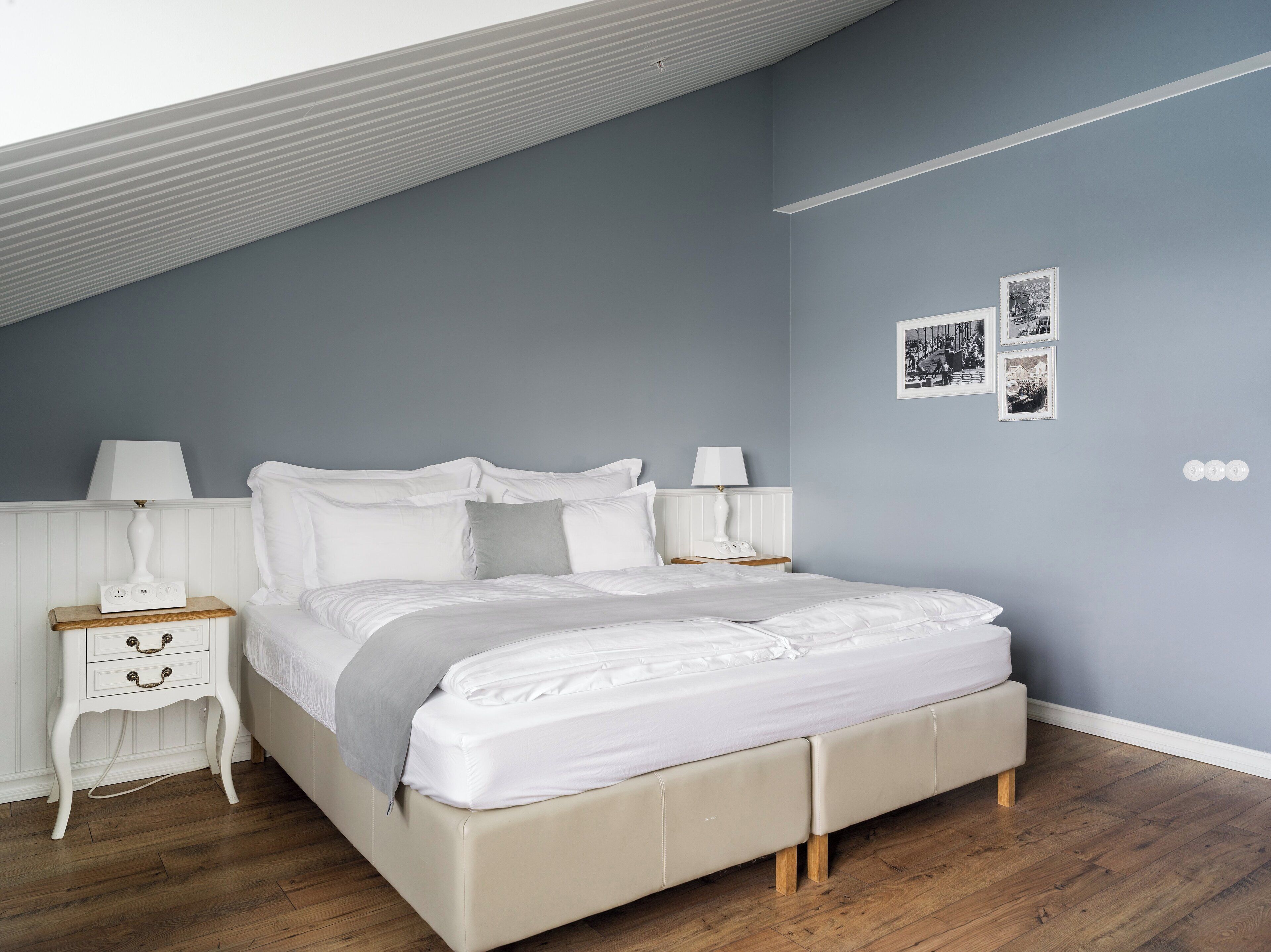 Une chambre avec un plafond en pente, un lit recouvert d’une literie blanche, deux tables de chevet avec des lampes et un mur orné de trois tableaux encadrés.
