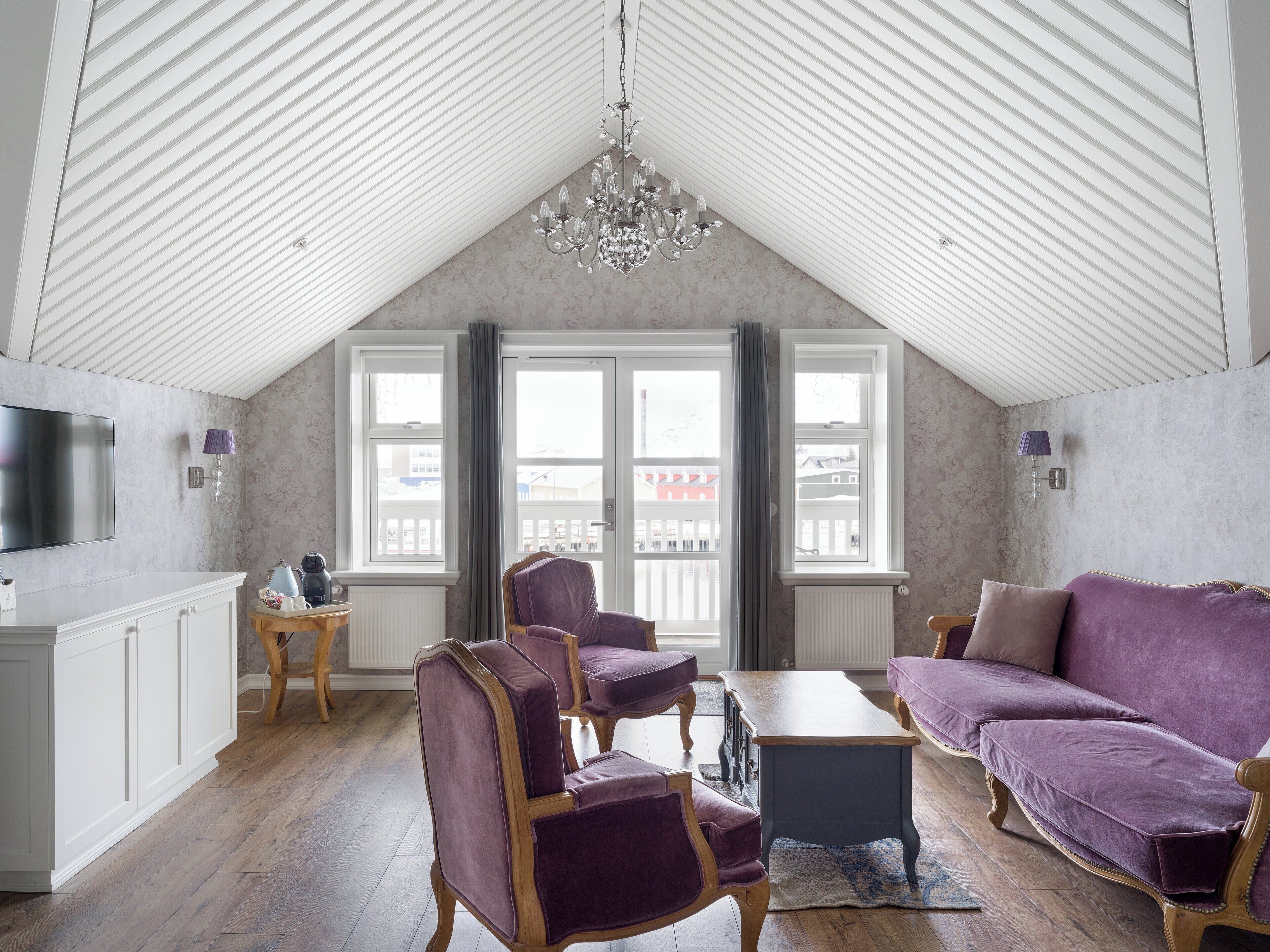 A spacious loft with a high ceiling, large windows, a modern chandelier, and a cozy seating area with purple upholstered furniture.