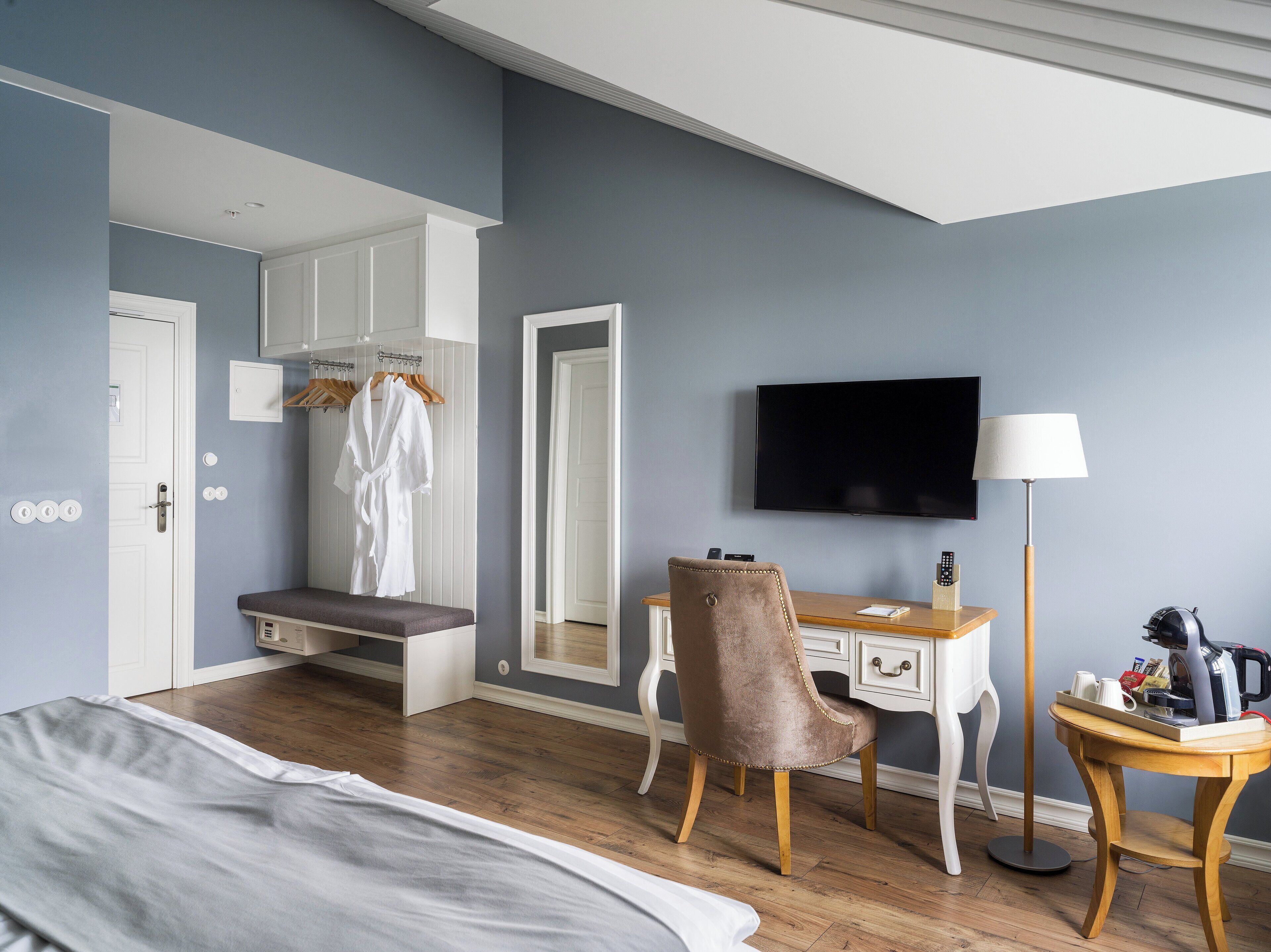 A hotel room with a bed, a desk with a chair, a TV, and a robe hanging on a rack.