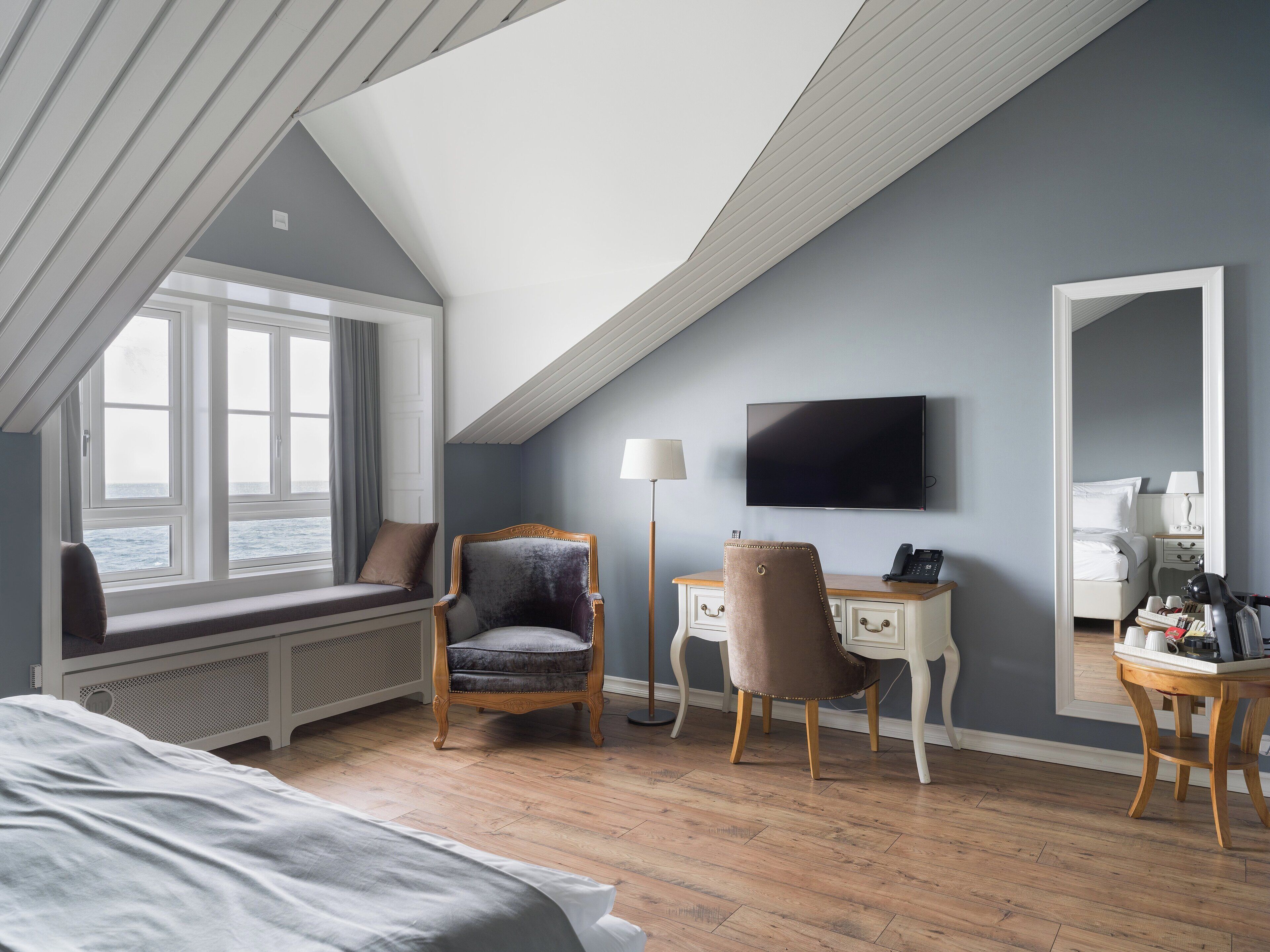 A hotel room with a sloped ceiling, a bed, a desk, a chair, a TV, and a window with a view of the sea.