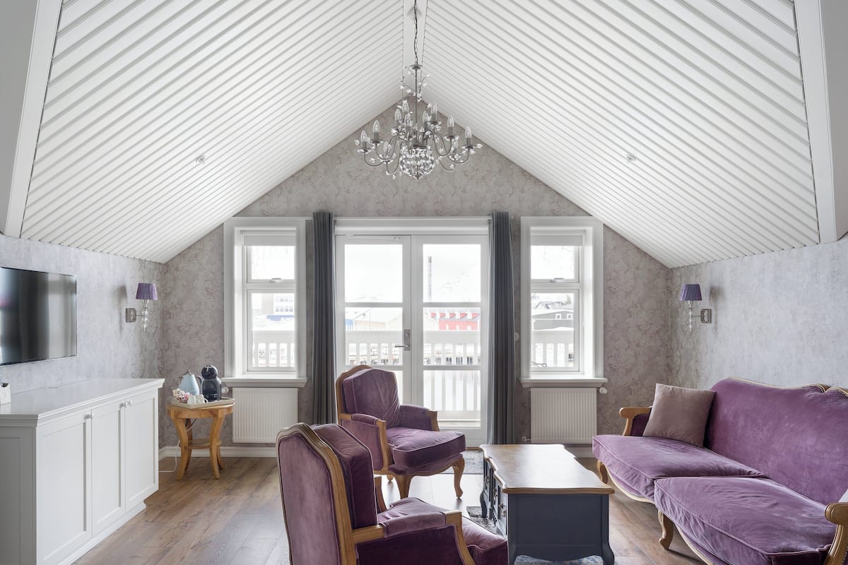 Un loft spacieux avec de hauts plafonds, de grandes fenêtres, un lustre moderne et un coin salon confortable agrémenté de meubles recouverts d’un tissu violet.