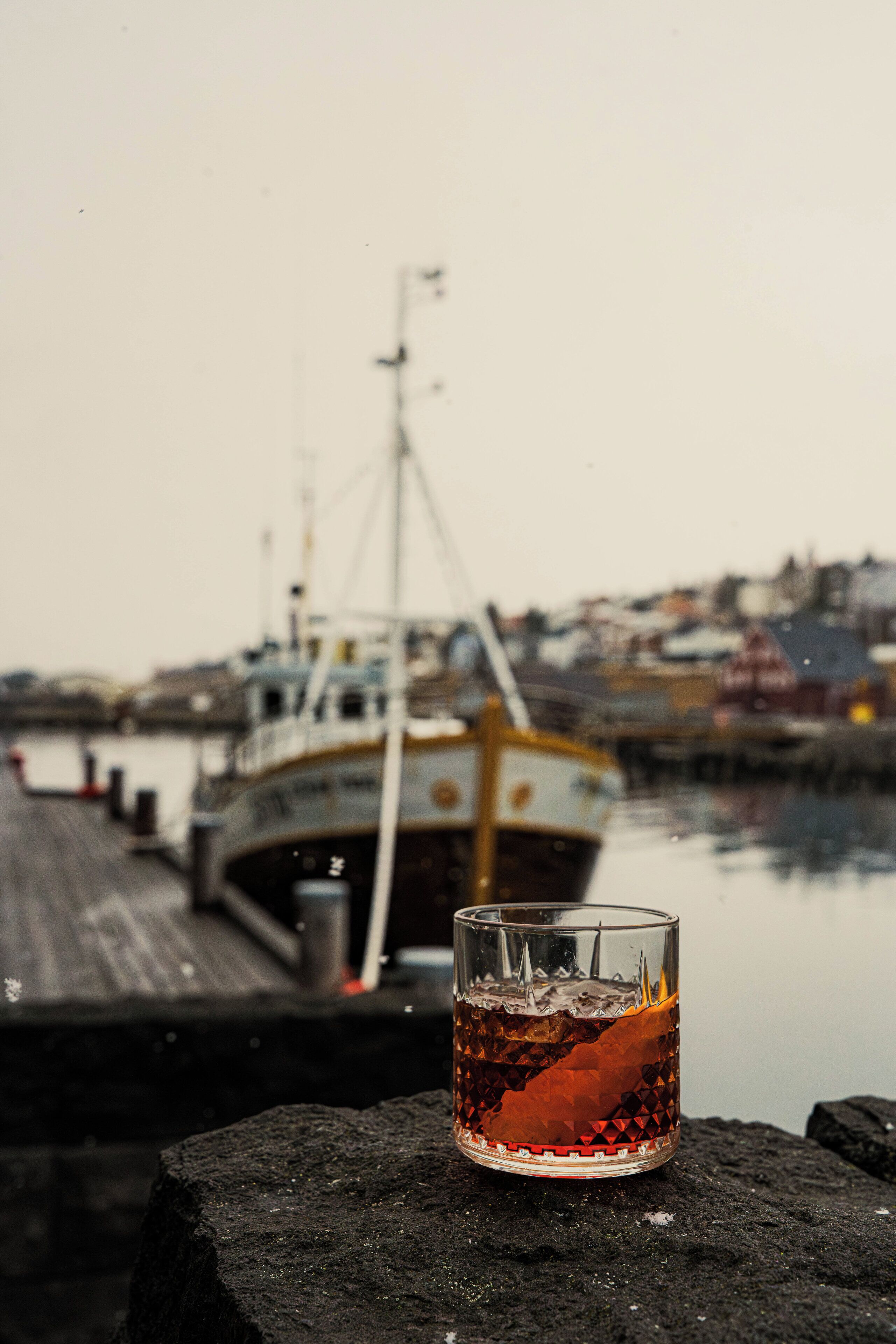 A glass of amber liquid on a dark surface with a harbor and boats in the background.