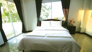 Una cama bien tendida en una habitación con un ventanal grande, una mesita de noche con un florero, y un suelo de baldosas.