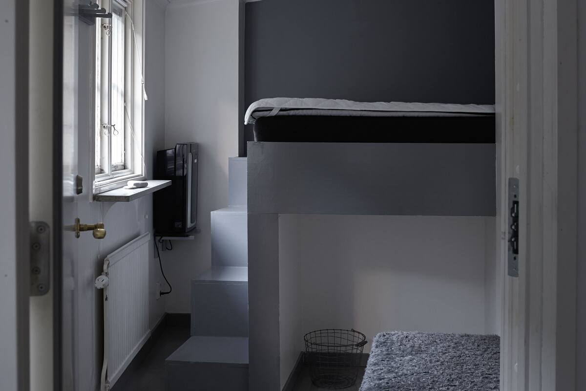 Een kleine kamer met een stapelbed, een klein raam, een radiator en een televisie.
