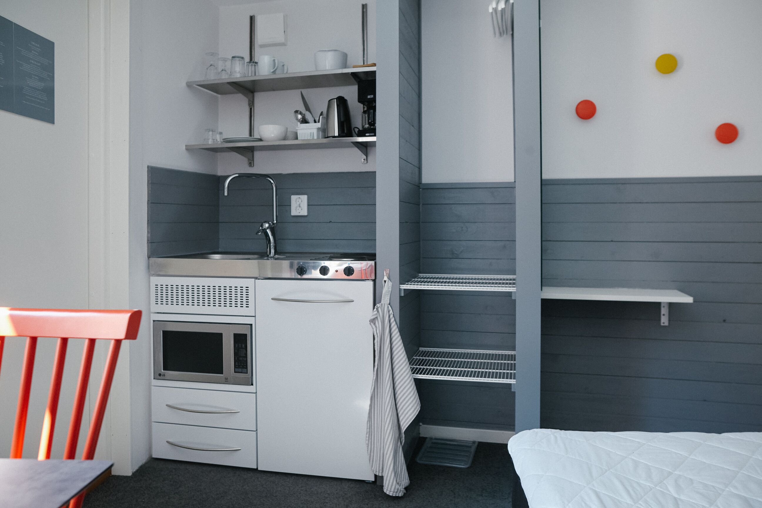 Una kitchenette compacta con microondas, horno y lavamanos, una silla roja y una cama con colcha blanca.